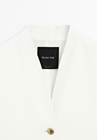 Blazer blanc avec un décolleté en V profond, fermeture à bouton doré unique et un tissu lisse et structuré. Étiquette de marque "Massimo Dutti" à l'intérieur.