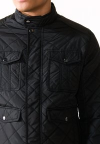 Black quilted jacket featuring a high collar, snap buttons, and four front pockets with flap closures. Smooth synthetic material and diamond pattern.
