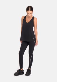 Melns sporta topiņš ar V-veida kakla izgriezumu un elegantu, brīvu piegriezumu, apvienots ar melnajām legingām, kuru sānos ir balta svītra, un melnām sporta kurpēm.