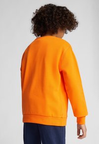 Sudadera de color naranja brillante con mangas largas, puños acanalados y cuello redondo. Tejido texturizado con un ajuste holgado, combinada con pantalones azul oscuro.