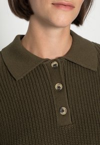 Tmavě olivově zelený pletený polo tričko s texturovaným povrchem, se stojáčkovým límcem a třemi béžovými knoflíky vpředu.