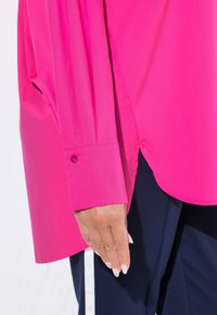 Fuchsia Langarmhemd mit Knopfmanschette, glatter Textur und seitlichem Schlitzdesign, kombiniert mit dunklen Hosen mit weißen Streifen.