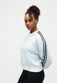 Jeune femme aux cheveux bouclés en chignon portant un sweat-shirt bleu clair avec le numéro "22" et un pantalon noir, debout devant un fond blanc.