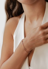 Pulsera de oro rosado con un delicado diseño de cadena, que presenta pequeñas cuentas y una barra curva lisa, llevada en un brazo desnudo.