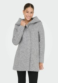 Femme souriante, portant un manteau à capuche gris clair en tissu texturé, un pantalon noir et des boucles d'oreilles en or, sur un fond clair et uni.