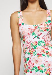 Žena v bezrukávových bílých šatech se vzorem růžových, červených a zelených květin, zobrazená v horní části těla a levé paži na jednoduchém pozadí.
