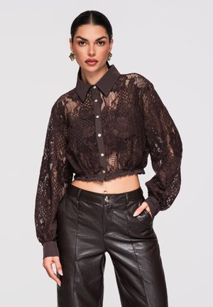 Kobieta w ciemnobrązowej przezroczystej koronkowej bluzce z krótkim rękawem, zapinanej na guziki z kołnierzykiem, oraz w czarnych skórzanych spodniach z wysokim stanem, z ręką w kieszeni.
