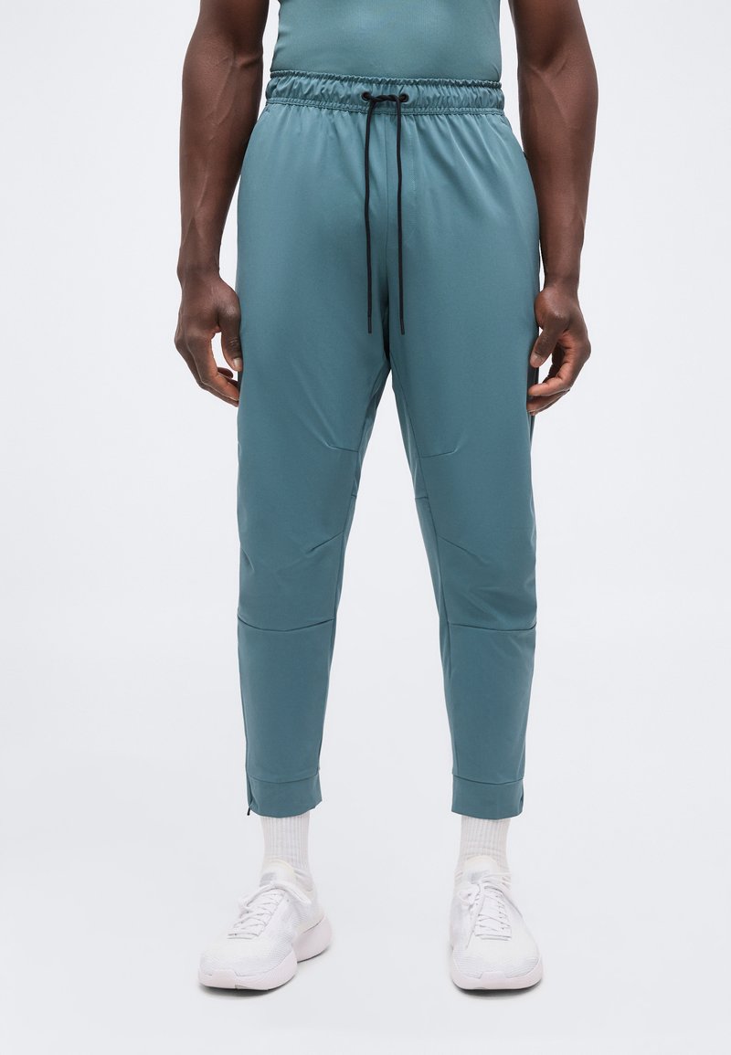 Άνδρας φοράει τυρκουάζ στενά αθλητικά παντελόνια με μαύρο κορδόνι, λευκές κάλτσες και λευκά αθλητικά παπούτσια, στέκεται σε απλό λευκό φόντο.