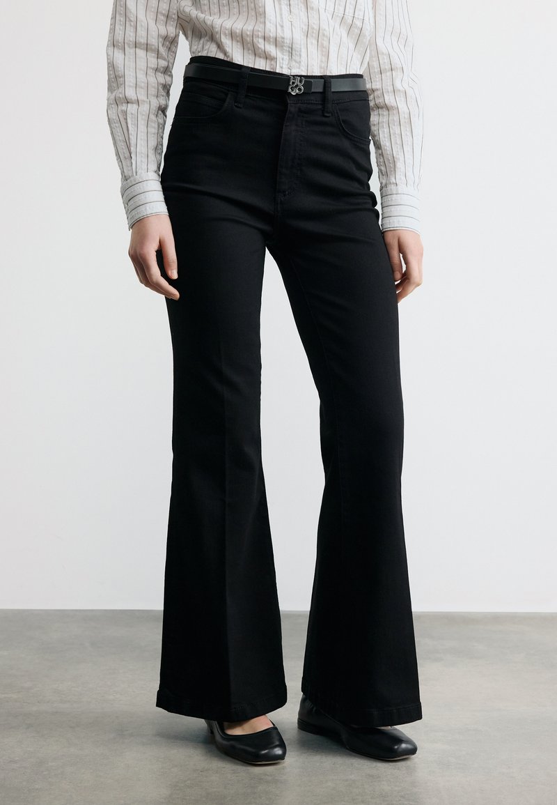 Ženska, oblečena v črne visokopasovne razširjene hlače, črni pas, črne baletne copate in belo dolgoročno majico z črtami in manšetami.