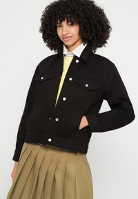 Črna denim jakna s srebrno opremo, s klasičnim ovratnikom, prsimi žepe in oprijeto zasnovo. Nosi se čez svetlo rumen pulover.