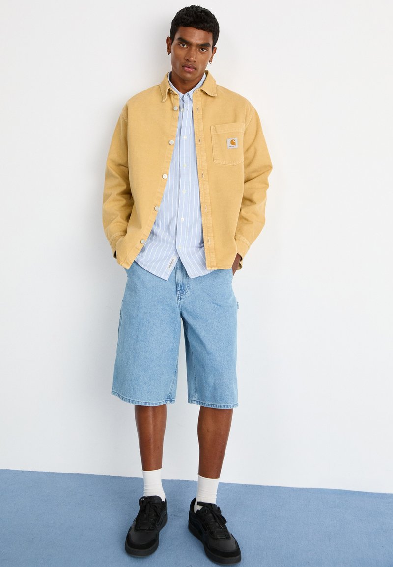 Veste en velours côtelé jaune sur une chemise à rayures bleues à boutons, associée à un short en denim bleu clair et des baskets noires avec des chaussettes blanches.