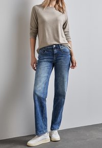 Jasnobeżowa bluzka z długim rękawem w połączeniu z niebieskimi jeansami o prostych nogawkach. Jeansy posiadają przednie kieszenie i luźny fason. Biały sneakersy dopełniają stylizację.