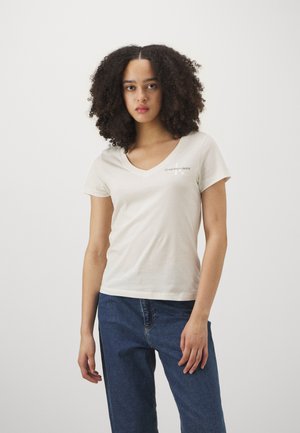 Beżowy t-shirt z dekoltem w serek, wykonany z miękkiej bawełny, z małym logo na lewej piersi. W połączeniu z niebieskimi dżinsami. Gładka tekstura i dopasowany krój.