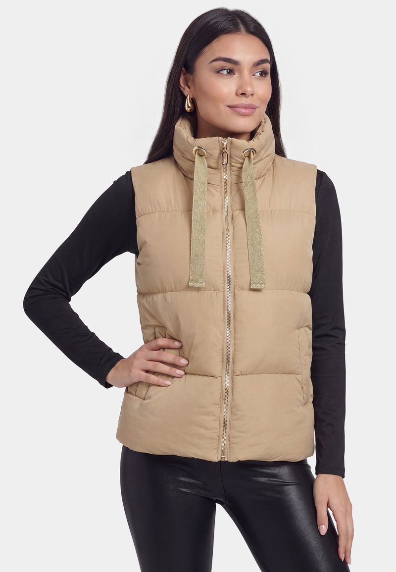 Mujer con cabello largo y oscuro lleva una chaqueta sin mangas acolchada color tostado con cordones sobre mangas largas negras y pantalones negros, de pie con la mano en la cadera.