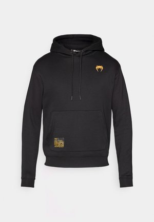 Schwarzer Hoodie aus weichem Stoff, mit einer Fronttasche, einer Kordelzugkapuze und einem goldenen Logo auf der Brust, das mit einem strukturierten gelben Etikett versehen ist.