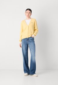 Žlutý cardigan s knoflíky, strukturovaný pletený, spárovaný se světle modrými širokými džíny a bílými teniskami, na neutrálním pozadí.