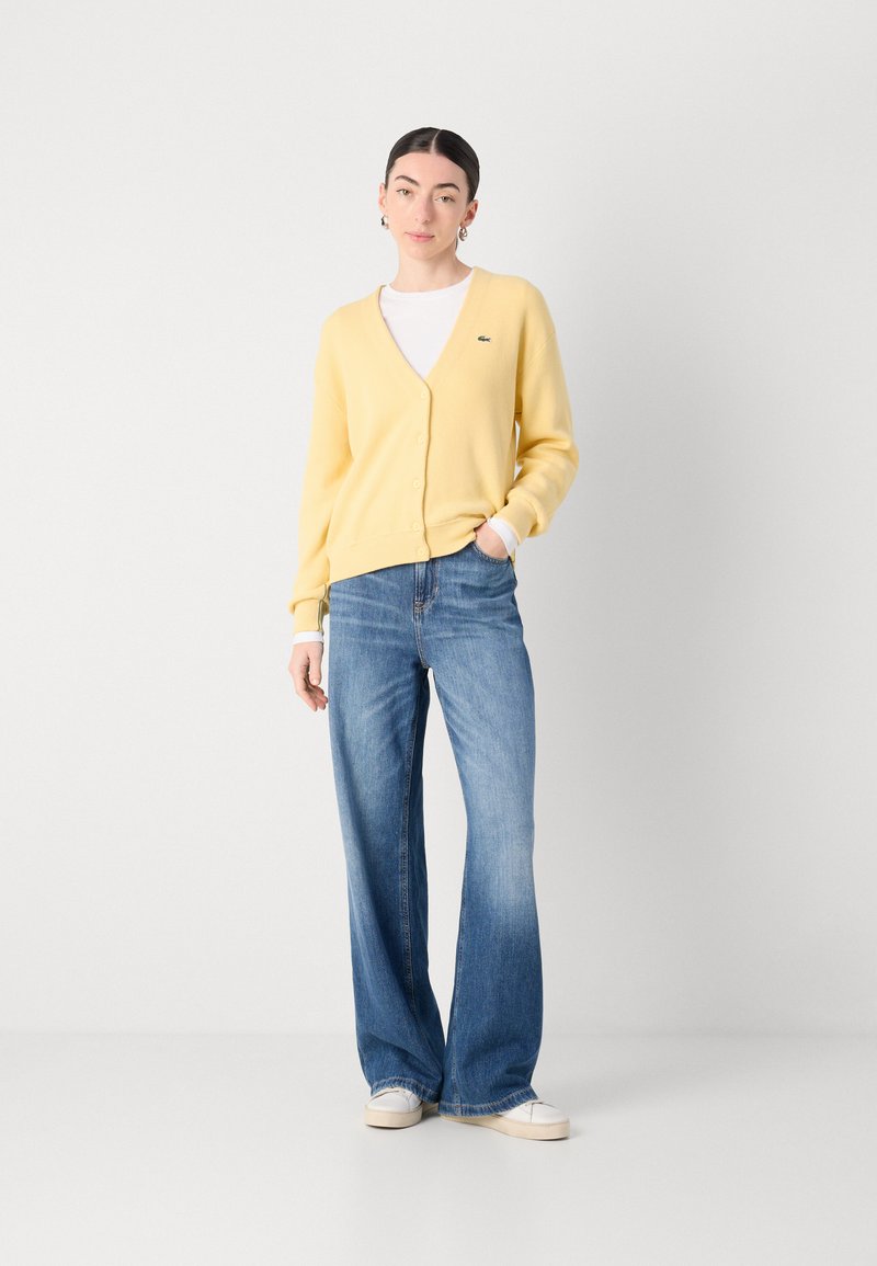 Gelbes Cardigan mit Knöpfen, strukturierter Strick, kombiniert mit hellblauen, weiten Jeans und weißen Sneakers, vor einem neutralen Hintergrund.