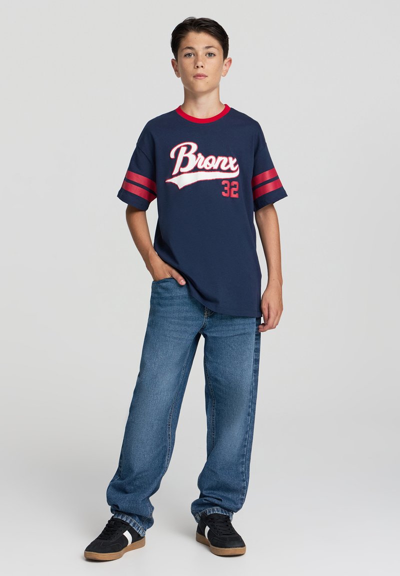 Granatowy t-shirt z czerwonymi akcentami, z białym napisem "Bronx 32", połączony z luźnymi niebieskimi dżinsami i czarnymi trampkami.