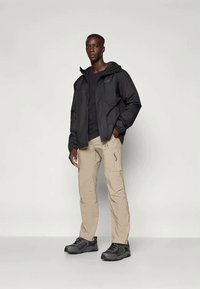 Jachetă neagră impermeabilă cu glugă, purtată peste o cămașă neagră, combinată cu pantaloni cargo bej și pantofi de drumeție negri. Design simplu, cu buzunare.