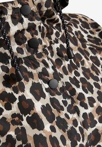 Leopardprintet stof i beige, brunt og sort, med sorte knapper og en snoretrækshætte til justerbar pasform. Glat tekstur.