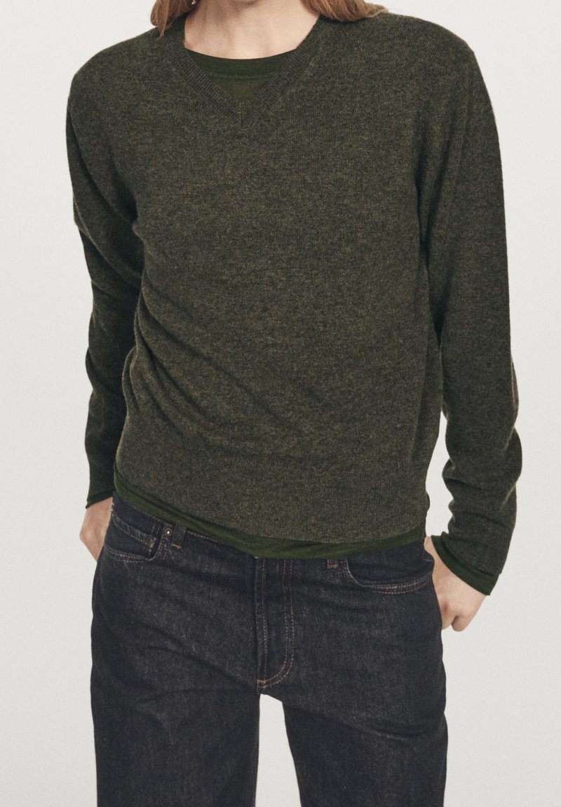 Personne portant un pull en maille vert foncé sur une chemise vert plus foncé et un jean bleu foncé, debout les mains dans les poches.