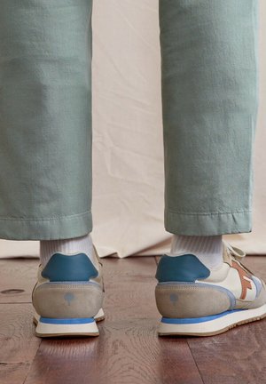 Personne portant des baskets beiges et bleues avec des chaussettes blanches et un pantalon vert clair, debout sur un sol en bois devant un fond beige.