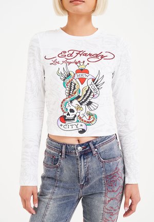 Witte lange mouwen crop top met kleurrijke tattoo-stijl graphic featuring een adelaar, slang en schedel; gecombineerd met blauwe denim jeans met rode accenten.