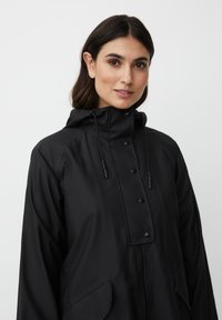 Giovane donna con capelli scuri che indossa una giacca impermeabile nera con cappuccio, dotata di bottoni automatici e cerniera, in piedi di fronte a uno sfondo bianco uniforme.