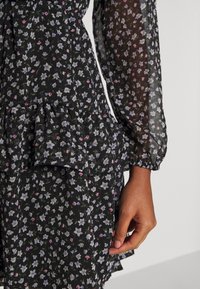 Schwarzes Blumenkleid aus semi-transparentem Material, mit langen Ärmeln, einem geschichteten Rüschensaum und kleinen lila und rosa Blumenmustern.