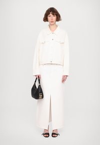 Giacca di jeans bianca con chiusure a bottone, abbinata a una lunga gonna bianca con uno spacco frontale. Borsa nera con la scritta "LOVE" in lettere dorate.