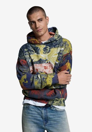 Uomo con capelli corti che indossa una felpa colorata con cappuccio con motivi di uccelli e jeans blu, braccia incrociate, guarda di lato.