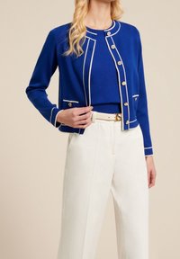 Stickad blå cardigan med vit kant, guldknappar och två framfickor, tillsammans med en blå topp och vita byxor.