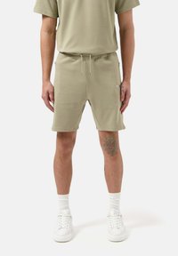 Pantaloni corti in cotone verde oliva chiaro con vita elasticizzata, cordino e tasche laterali. Indossati con scarpe da ginnastica bianche e calze.