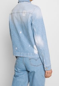 Světlomodrá džínová bunda s vybledlými skvrnami a opotřebením. Dlouhé rukávy, klasický límec, zapínání na knoflíky a minimalistické designové detaily.