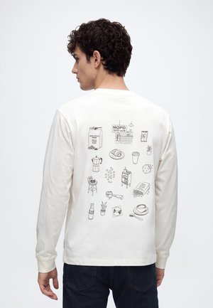 Homme portant une chemise blanche à manches longues avec des dessins au trait noirs d’objets de café et de plantes dans le dos sur un fond uni.