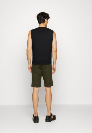 Mann trägt ein schwarzes ärmelloses Shirt, olivgrüne knielange Shorts und schwarze Sandalen, steht vor einer schlichten weißen Wand.