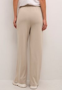 Pantalon beige à jambes larges en tissu doux avec une ceinture élastique, présentant une texture lisse et une coupe décontractée, associé à des chaussures blanches.