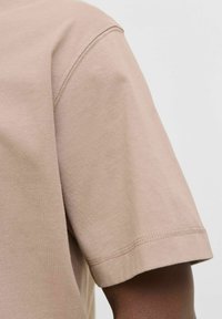 Beige katoenen T-shirt met een ronde hals en korte mouwen, met zichtbare stiksels langs de zoom en mouwranden.