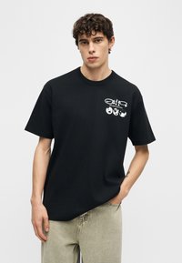 Jonge man die een zwart T-shirt draagt met witte tekst 'Nature in Motion' en pictogrammen van de globe, beige broek, staand met één hand in de zak.