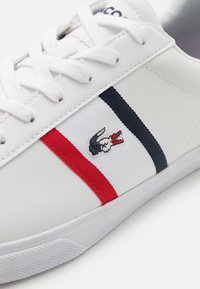 Adidași albi cu finisaj texturat, având accente în dungi albastre și roșii și un logo brodat de crocodil pe lateral.