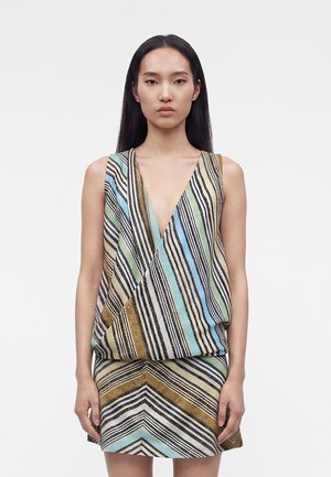 Egyenes fekete hajú nő, aki ujjatlan, színes csíkos ruhát visel mély V-kivágással, egyszínű fehér háttér előtt áll.