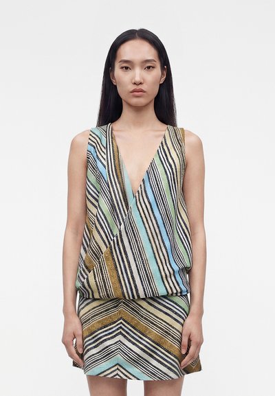 Femme aux cheveux noirs lisses portant une robe sans manches multicolore à rayures avec un profond décolleté en V, debout devant un fond blanc uni.