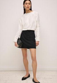Top blanco de manga larga con cuello redondo y mangas ligeramente acampanadas, combinado con una falda mini negra. Bolso de mano negro y zapatos planos con tachuelas.