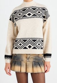 Personne portant un pull beige avec des motifs géométriques noirs et une mini-jupe plissée en denim délavé avec des collants noirs.