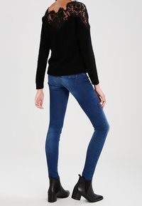 Schwarzer Pullover mit Spitzendetail am Rücken, kombiniert mit blauen Skinny-Jeans und schwarzen Stiefeletten. Weiche Textur, figurbetonte Silhouette und lässiger Stil.