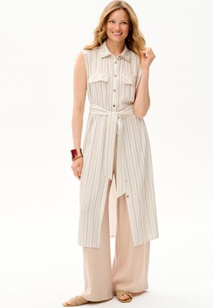 Uśmiechnięta kobieta ubrana w bezrękawową, paskowaną sukienkę koszulową z paskiem, na szerokich beżowych spodniach i brązowych sandałach, z dodatkami w postaci czerwonej bransoletki i kolczyków.