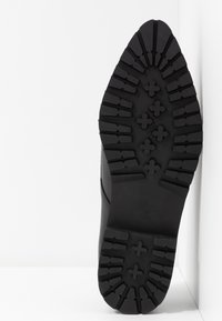 Semelle en caoutchouc noir d'une chaussure, présentant un motif de semelle texturé avec des rainures profondes et des formes croisées surélevées, conçue pour l'adhérence. Taille 37.