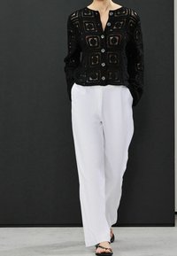 Schwarze Häkeljacke mit quadratischen Mustern, runden Knöpfen und langen Ärmeln, kombiniert mit weißen, locker sitzenden Hosen. Flache Sandalen mit Riemen.