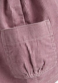 Tejido de pana rosa con costillas verticales, que presenta un bolsillo patch cuadrado y un detalle fruncido en el dobladillo. Textura suave y color apagado.