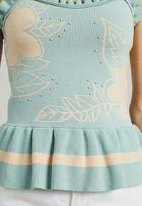 Jasnoniebieski dzianinowy top z kwiatowymi wzorami w kolorze kremowym, małymi teksturowanymi kropkami oraz rozkloszowanym dołem z paskowym akcentem w kolorze kremowym i brzoskwiniowym.